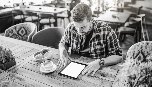 英俊的白人男子在格子衬衫手在桌子上看平板电脑。桌子上的耳机和咖啡，咖啡厅内部。黑白照片。图片下载