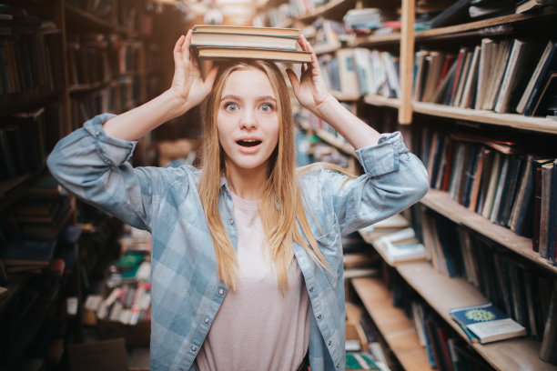 一个女孩站在书架旁边的有趣图片。她双手顶着两本书，眼睛直视前方。她看起来很惊讶。图片下载