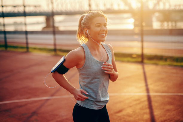 迷人的金发女跑步者穿着运动服在球场上跑步，耳朵里戴着耳机，胳膊上挎着手机套里的智能手机。早上健身的概念。跑得慢不是性格缺陷，放弃才是。图片下载