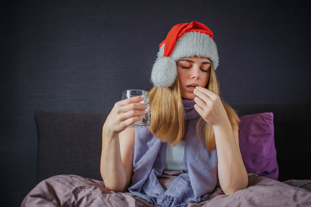 一张年轻女子坐在床上吃药的照片。她用另一只手盛着水。她手上戴着圣诞帽，脖子上围着围巾。图片下载