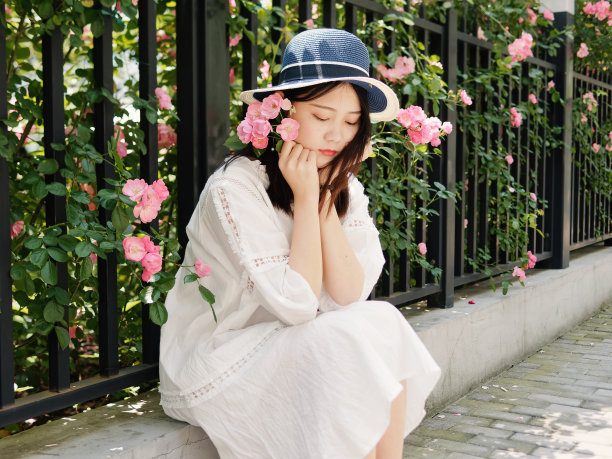 户外肖像美丽的中国妇女在白色的裙子微笑在粉红色的玫瑰墙花园，快乐的女孩享受她的闲暇时间。图片下载