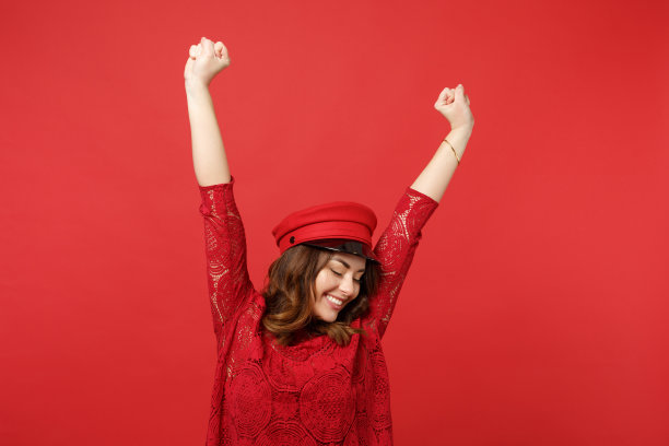 快乐的年轻女子在蕾丝连衣裙，帽子保持闭上眼睛紧握的拳头，举起的手孤立在明亮的红色墙壁背景在工作室。人真诚情感，生活理念。模拟拷贝空间。图片下载