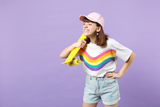 微笑少女身穿亮色衣服手持黄色滑板的写真，在紫色背景中拍摄。人物真挚的情感，生活方式概念样机图片下载
