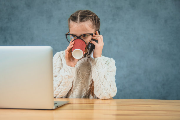 快乐的小女孩正坐在桌旁，拿着她的灰色笔记本电脑和一部智能手机，孤立在灰色的背景中。图片下载