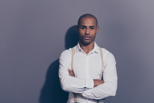 特写照片美丽惊人的黑皮肤，他，他，他的大男子主义的胳膊，折叠着看直相机，善良真诚的眼睛，闪亮的剃须的脸，穿白衬衫，柔和的吊带，衣服孤立的灰色背景图片下载