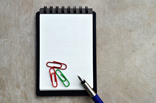 一张水平的照片，上面是一个普通的空白白纸，一个螺旋装订的垂直记事本，黑色封面，一只蓝色和银色的金属钢笔，顶部放着三个回形针，背景是木质的米色，陈旧的古董图片下载