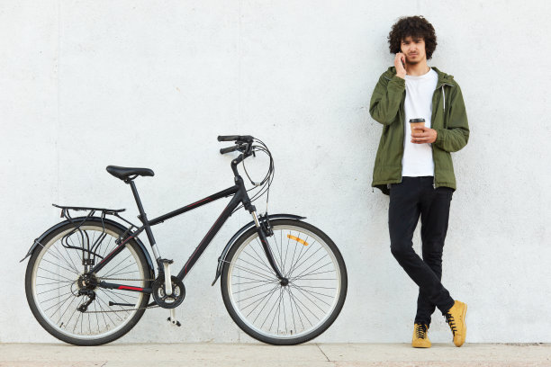 一个卷发帅哥的形象，穿着绿色的夹克，黑色的裤子，留着卷发，和别人通过手机聊天，在自行车旁边喝着外卖咖啡，准备骑车。图片下载