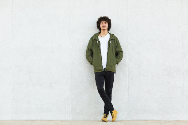 这张照片拍摄的是一个长着小胡子和卷曲的黑色头发的欧洲男性，穿着时尚的夹克，在白色集中的墙壁上孤立，直视着镜头，脸上带着快乐的表情。图片下载