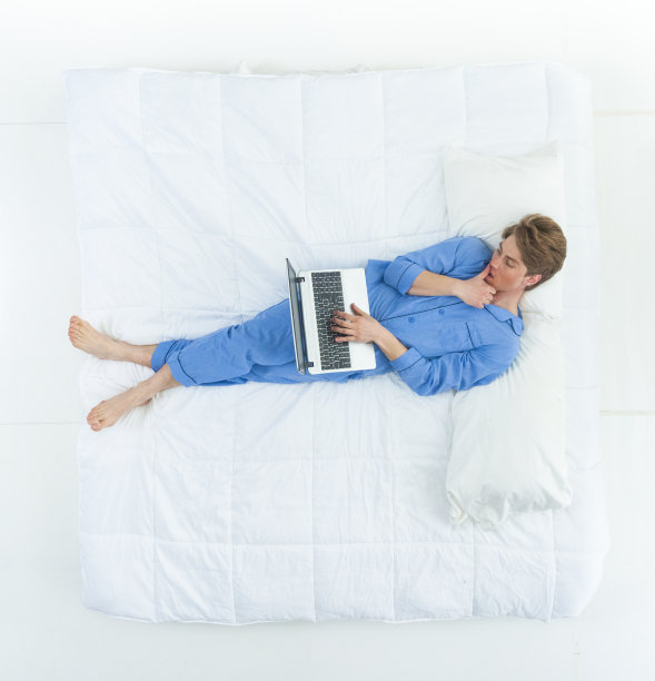 英俊的男人穿着睡衣躺在床上用着他的工作笔记本电脑图片下载