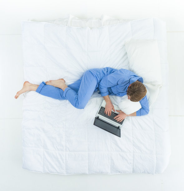 英俊的男人穿着睡衣躺在床上用着他的工作笔记本电脑图片下载