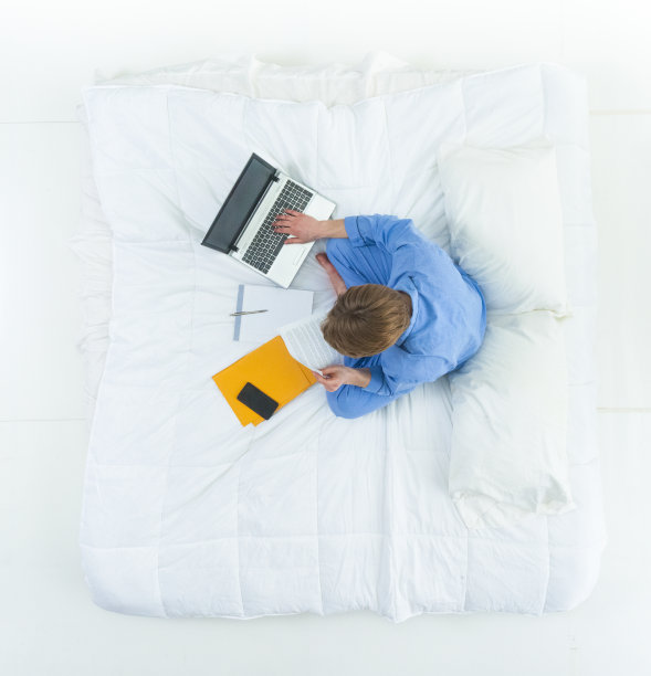 英俊的男人穿着睡衣躺在床上用他的工作笔记本电脑和文件图片下载