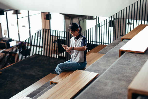 一名亚洲女性坐在楼梯上，一边看手表一边用智能手机读短信、拍照片——教育和商业理念图片下载