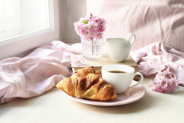 春天的静物景象。一杯咖啡，牛角包，旧书和牛奶罐。复古女性风格的照片。花卉构图以粉红色的樱花，白色的樱花花桌上。图片下载