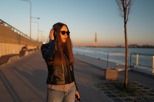 一名年轻女子穿着皮夹克和牛仔裤，戴着耳机通过手机听音乐，在道加瓦河附近的日落图片下载