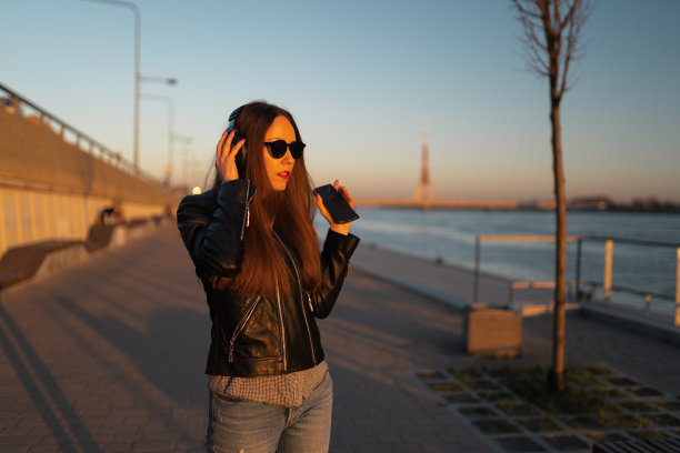一名年轻女子穿着皮夹克和牛仔裤，戴着耳机通过手机听音乐，在道加瓦河附近的日落图片下载