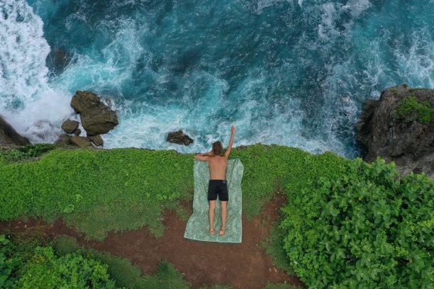 在印度尼西亚巴厘岛的乌鲁瓦图，一个人躺在蓝色的印度洋上的绿色悬崖上，浪花翻滚。航拍照片，一名男子躺在蓝色的印度洋上的绿色悬崖上，浪花翻腾，印度尼西亚巴厘岛的乌鲁瓦图。图片下载