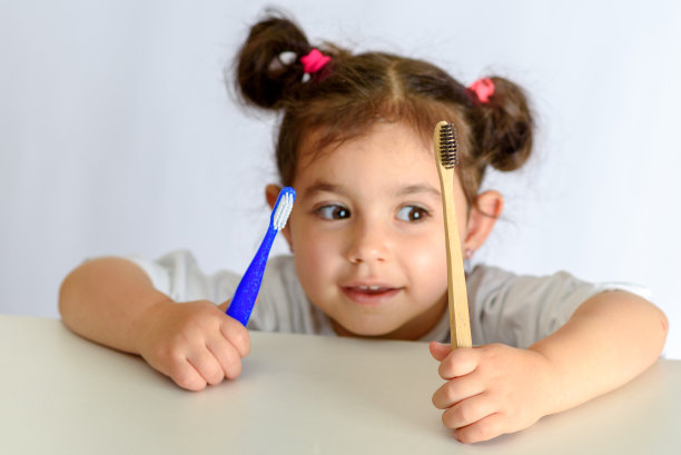 小女孩穿着白衬衫，拿着竹牙刷和塑料牙刷。图片下载