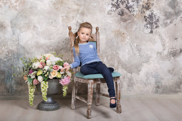 一个迷人的微笑小女孩的肖像穿着蓝色的毛衣和裤子，她的头发叠在她的头发坐在椅子上，对着一个垃圾墙的背景。图片下载