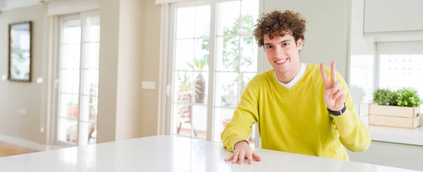 广角拍摄的年轻英俊的男子在家里微笑看着镜头显示手指做胜利的手势。二号人物。图片下载