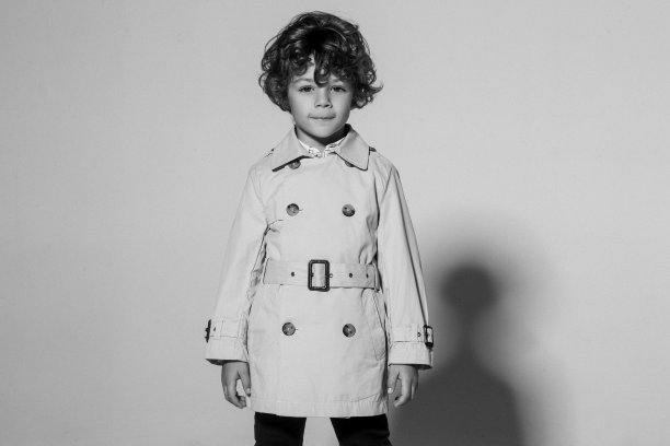 一个可爱的卷发小男孩优雅的肖像，穿着雨衣摆出正面姿势，孤立在一个白色的背景。黑白照片。图片下载