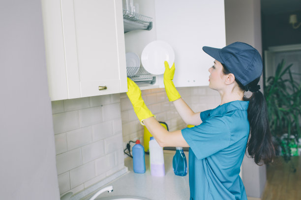 清洁工把盘子放在碗柜里的图片。它被打开。女孩穿着蓝色的制服和黄色的手套。她清理空间。图片下载