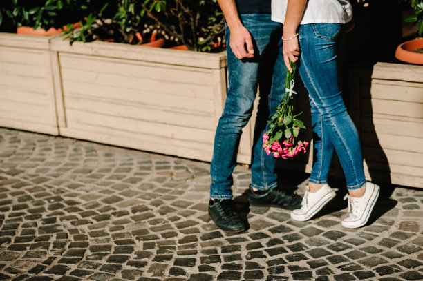 这张照片里的女人和男人穿着牛仔裤和鞋子，女孩捧着一束花踮着脚尖，时髦的情侣在约会中接吻。户外爱好者。图片下载