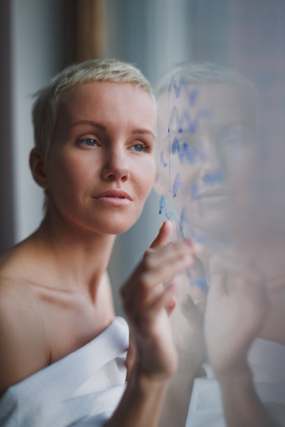 美丽的年轻女子，一头金色短发，站在窗前，画出蓝色的海浪。垂直的照片。图片下载