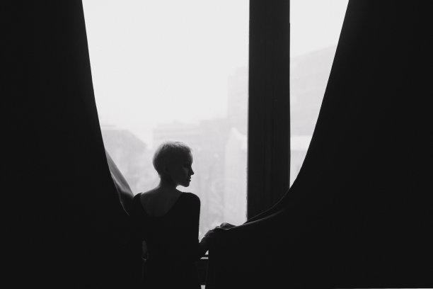 在一间挂着深色窗帘的房间里，站在窗前的是一位漂亮、苗条、留着浅色短发的年轻女子。黑白照片。图片下载