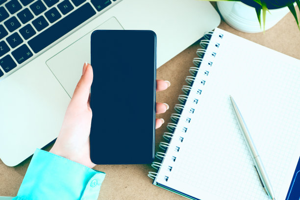 女性手握黑色手机智能手机与黑屏笔记本电脑和记事本背景。图片下载