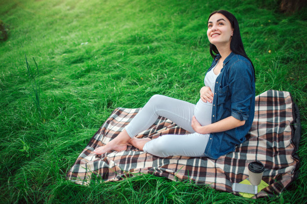一个快乐的黑发和骄傲的孕妇肖像在一个城市的公园里。照片中的女模特用手摸她的肚子。女模特正坐在草地上。图片下载