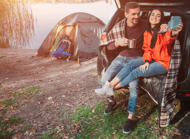 一名年轻女子和一名男子坐在后备箱里，对着镜头摆姿势。她有问题。年轻女人手里拿着它。家伙杯。他们在湖边。图片下载