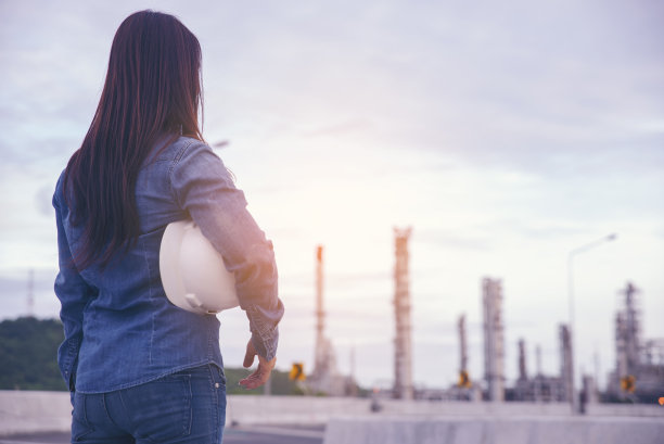 女工程师和安全主任的概念。年轻的女工程师手持安全帽(白色安全帽)，检查员在炼油厂背景前。图片下载