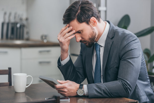 一个悲伤的男人穿着灰色西装坐在木桌上，拿着智能手机，白色的杯子，看着相框里的照片，在家里思考，悲伤的混乱概念图片下载
