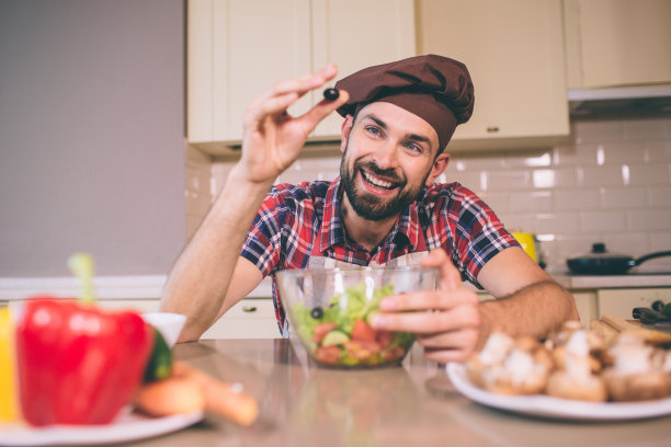 一个男人站着，靠在桌子上的漂亮照片。他一只手拿着黑橄榄，另一只手放在碗里。男人看起来很开心，很满足。图片下载