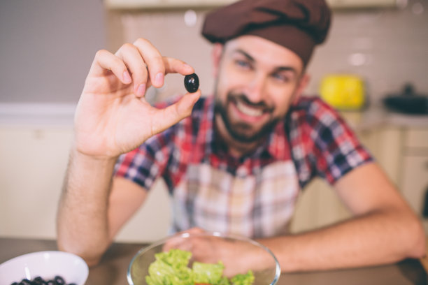 一张正面的、蓄着胡须的男人倚靠在桌子上，手里拿着一块黑橄榄的照片。他看着它。年轻人专注于此。他把它展示给摄像机。图片下载