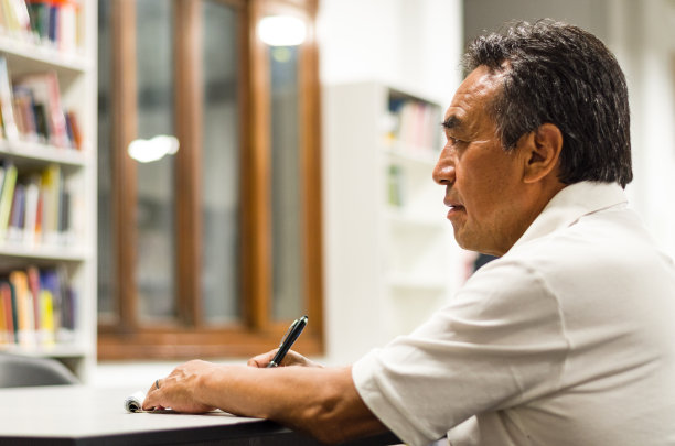 一位严肃的长者坐在图书馆的长椅上，在写他的书。图片下载