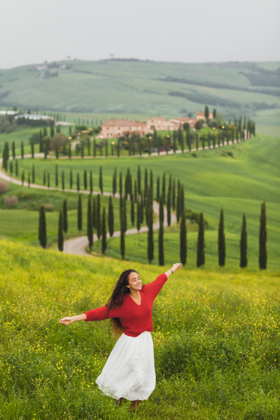 在托斯卡纳春天的风景背景上，穿着红色毛衣和白色裙子的女人图片下载