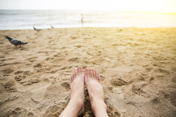 男人光着脚在海滩上放松。那家伙的腿在沙滩上。海浪在海边。在云和阳光下的海岸线。暑假。旅行的概念。图片下载