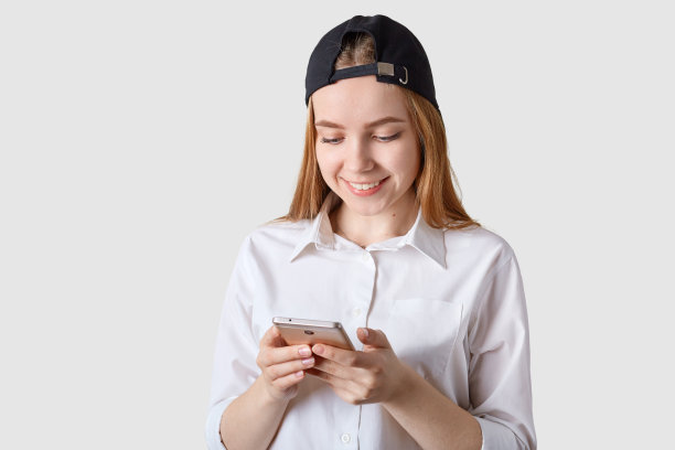 近距离的微笑美丽的少女的肖像与帽檐后，有棕色的头发，在白色的工作室墙壁，手持智能手机，查看社交网络，与朋友交流，穿着白色衬衫。图片下载