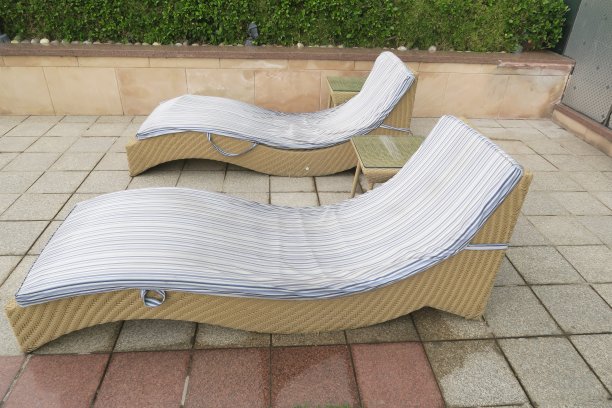 两个相邻的弯曲的柳条太阳躺椅/太阳躺椅/蒸汽机在花园庭院的铺路板上游泳池的图像，雨后在院子外面的湿躺椅上有蓝白条纹的垫子，旁边的花园树篱照片图片下载