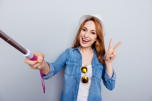 近距离的照片美丽的她她的女士旅行者手手臂金属自拍杆博主使照片v标志走在街上有趣的时髦的戴眼镜帽子休闲牛仔裤牛仔衬衫孤立的灰色背景图片下载