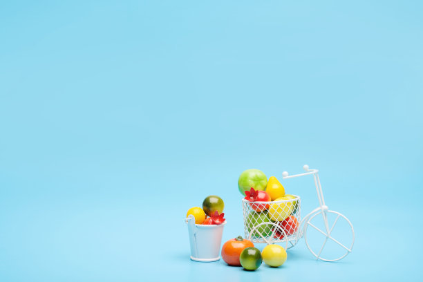白色钢丝自行车和一个装满迷你水果的篮子。靠近装着蔬菜和水果的桶。蓝色背景。收获和运送果蔬的理念。图片下载
