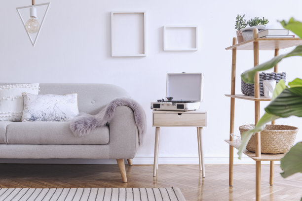 时尚舒适的斯堪的纳维亚白色内饰设计沙发，书架与配件，枕头，毯子，留声机和模拟相框。白色的背景墙和现代的三角形灯。图片下载