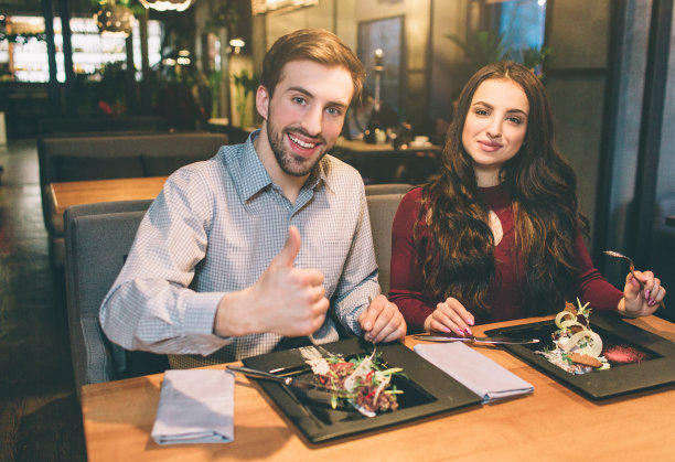 广告图片上的男人和女人坐在桌子上有一些食物。他们看着镜头微笑着。男人在竖起他的大拇指。图片下载
