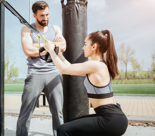 一个苗条女孩做下蹲的垂直图片。她拿着线圈以保持平衡。年轻男子站在她旁边看着女孩。图片下载