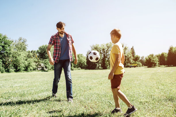 一张父子在踢足球的照片。有一个球从爸爸那里飞到儿子那里。它在空气中。外面是阳光明媚的球场。图片下载