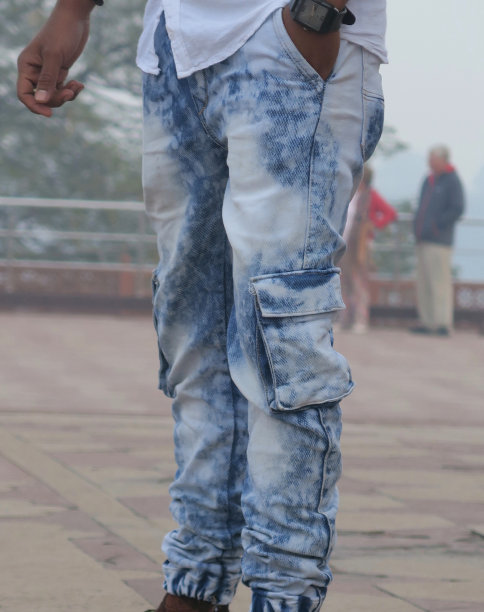 在模糊的天空背景和印度德里的游客映衬下，年轻的亚洲印度男子的形象，时髦男子的6个口袋的石洗漂白蓝色牛仔牛仔裤，松紧的牛仔裤脚照片和漂白粉图案的斑驳牛仔裤图片下载