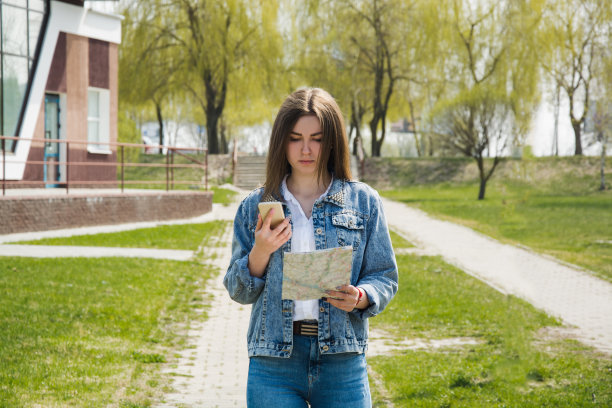 一个年轻的女孩拿着手机和地图正在寻找道路。女孩游客可爱的穿着牛仔裤的衣服走在街上在夏天，看地图和在路上。图片下载
