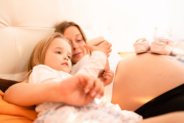 怀孕的母亲和年幼的女儿小孩子躺在床上玩智能手机看家庭照片自拍图片下载