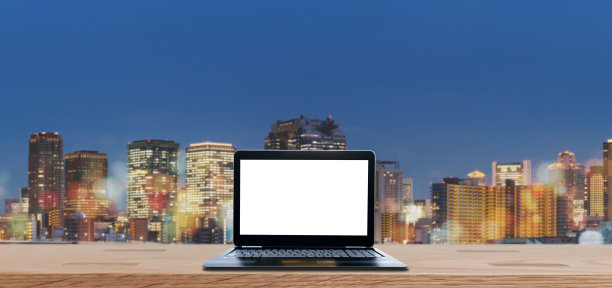 电脑笔记本电脑空白的白色屏幕上的木桌模糊了大阪城市的夜景在日本。笔记本电脑的背景图片下载